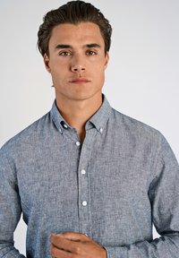 Camisa de botón en tejido chambray de color azul claro, con cuello, mangas largas y detalles de botones. Textura suave con sutiles variaciones.