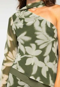 Blusa verde de un solo hombro hecha de tela ligera, con un estampado floral en tonos verdes y beige, y un escote drapeado.