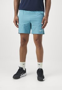 Pantalones cortos deportivos de color azul claro con bolsillos laterales, acentos negros y un logo de Nike blanco, combinados con zapatillas negras y calcetines de color claro.