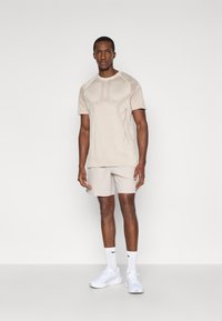 Μπεζ αθλητικό μπλουζάκι με υφή στους ώμους, συνδυασμένο με ανοιχτό μπεζ σορτς. Περιλαμβάνει κοντά μανίκια και στρογγυλή λαιμόκοψη.