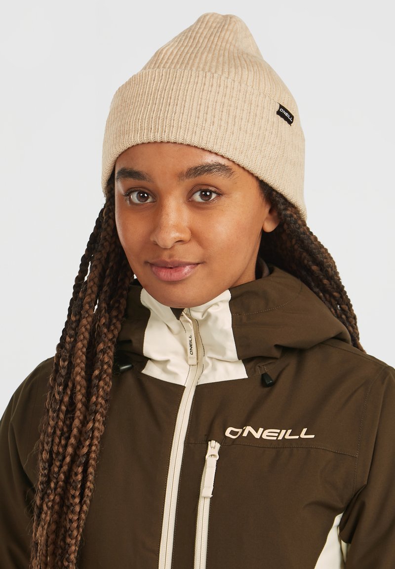 Beige strikket rib hat med en spids i toppen. Båret med en brun yderjakke med hvid lynlås og logo-detalje.