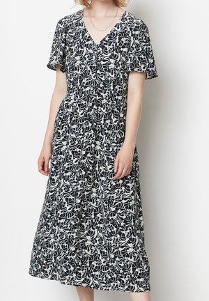 Žena v černo-bílých květovaných midi šatech s krátkými rukávy, knoflíky a výstřihem do V na jednobarevném bílém pozadí.