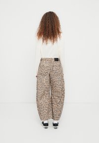 Άτομο με μακριά σγουρά μαλλιά, ντυμένο με λευκό μακρυμάνικο πουκάμισο, φαρδιά παντελόνια με λεοπάρ μοτίβο, λευκές κάλτσες και μαύρα αθλητικά παπούτσια, στέκεται μπροστά σε έναν λευκό τοίχο.