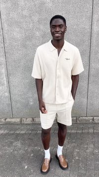 Beige knappet skjorte med kort erme og strukturert stoff, kombinert med lyse shorts og brune loafers. Hvite sokker fremhever fottøyet.