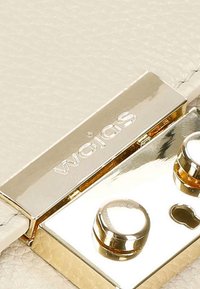 Krémová kožená taška v detailu s kovovou zlatou sponou s vyrytým nápisem „Wojas“, raženými akcenty a strukturovaným povrchem.