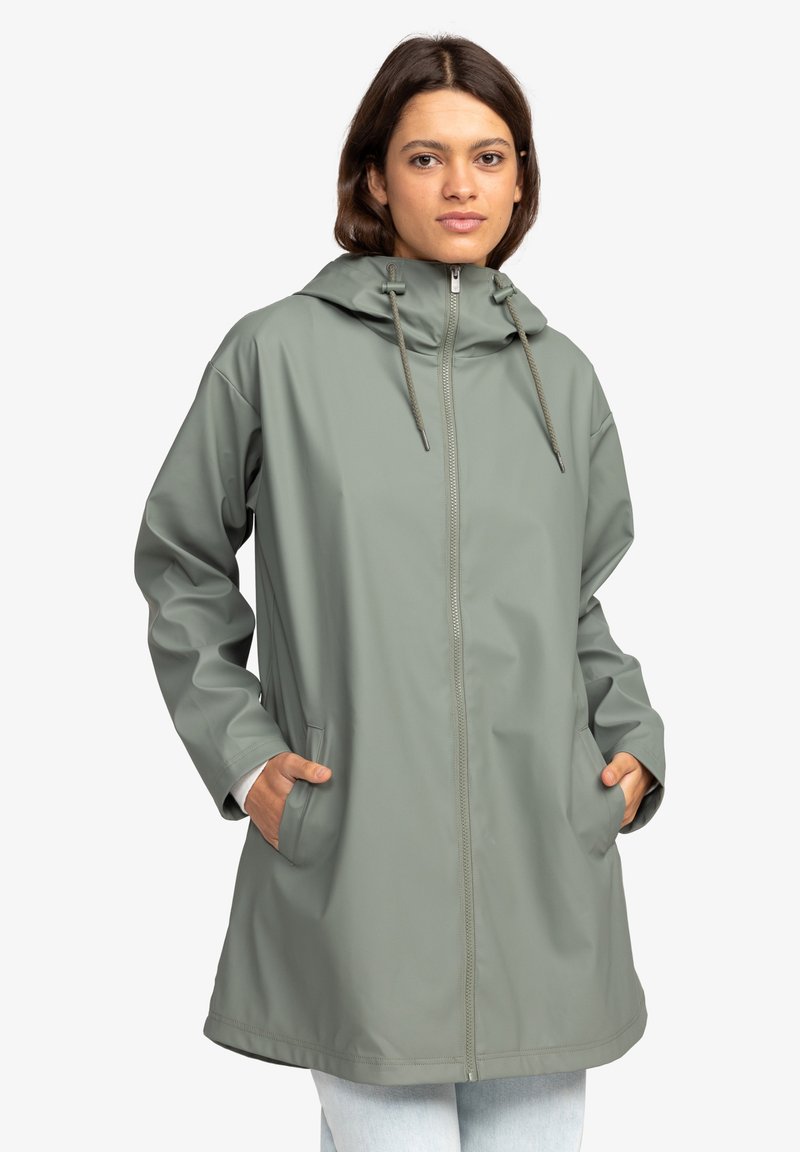 Capa de lluvia verde claro, impermeable, con capucha, cremallera frontal, cordón y bolsillos laterales, que presenta un ajuste relajado y una textura suave.
