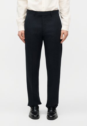 Mężczyzna ubrany w czarne formalne spodnie, czarne skórzane eleganckie buty oraz białą, długorękawową koszulę na guziki, stojący na tle jednolitego tła.