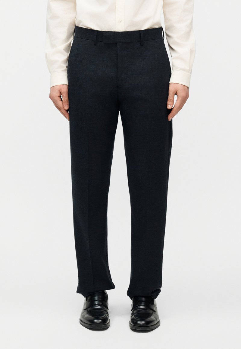 Άνδρας που φοράει μαύρο επίσημο παντελόνι, μαύρα δερμάτινα παπούτσια και λευκό πουκάμισο με μακριά μανίκια και κουμπιά, στέκεται μπροστά σε απλό φόντο.