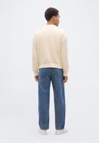 Homme debout, de dos, portant un pull crème, un jean large bleu avec des poches arrière, et des baskets blanches sur un fond blanc uni.