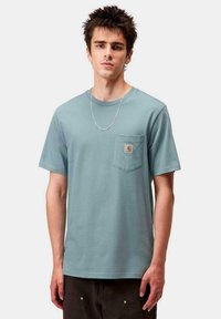 Joven con cabello de punta que lleva una camiseta Carhartt azul claro con bolsillo en el pecho y un delgado collar de plata, posando frente a un fondo liso.