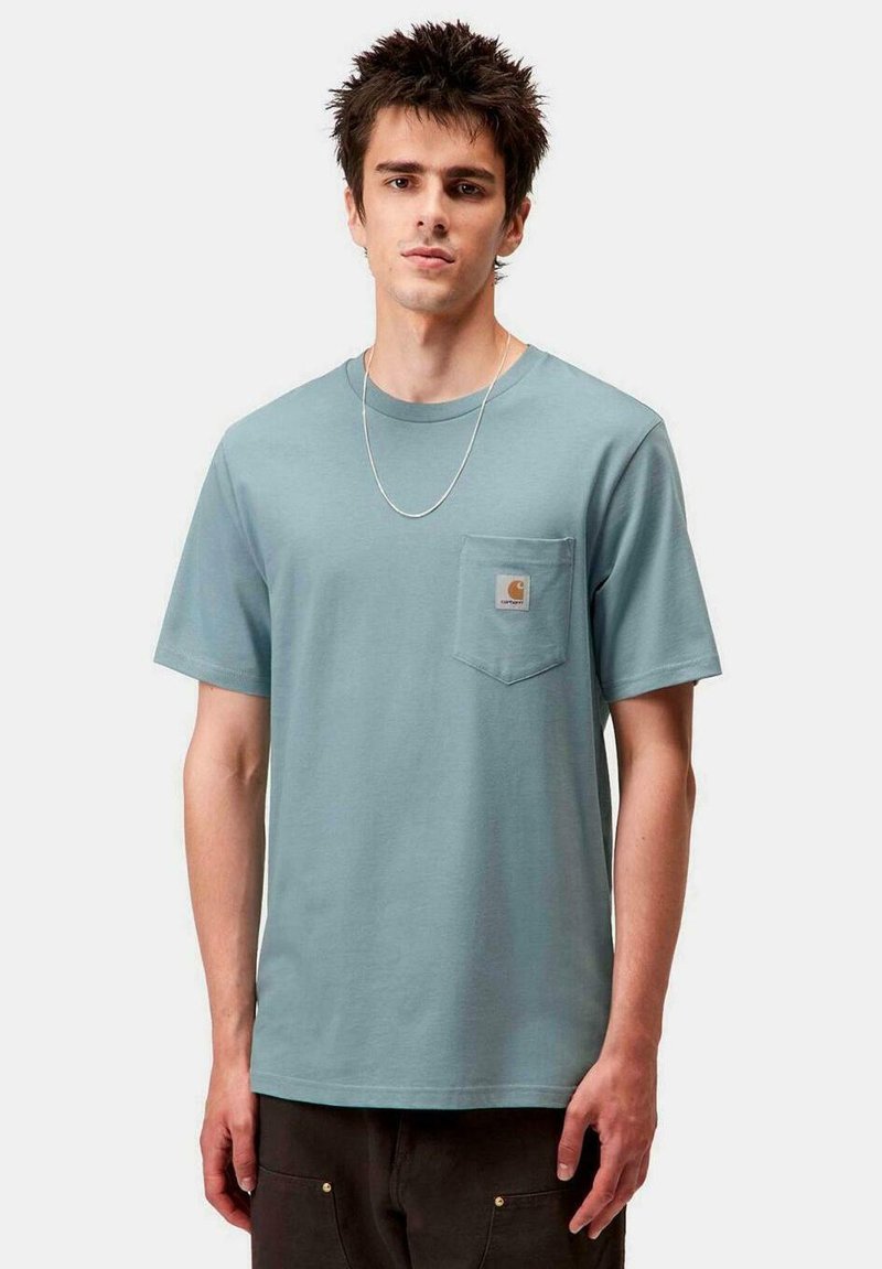 Joven con cabello de punta que lleva una camiseta Carhartt azul claro con bolsillo en el pecho y un delgado collar de plata, posando frente a un fondo liso.