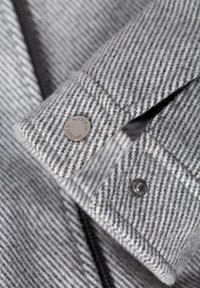 Grau gestreifter Stoff mit weicher Textur, der mit einem Druckknopfdetail versehen ist. Kragen und Manschetten zeigen ein ordentliches Finish und betonen die Handwerkskunst.