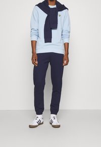 Helles blaues Sweatshirt mit gerippten Bündchen, marineblaue Jogginghosen und ein marineblauer Pullover um den Hals gebunden. Weiße Sneaker mit schwarzen Streifen runden den Look ab.
