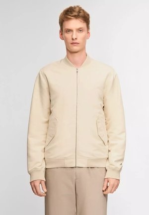 Jonge man staat, draagt een beige jas met ritssluiting en klepzakken en bijpassende lichtbruine broek tegen een effen achtergrond.