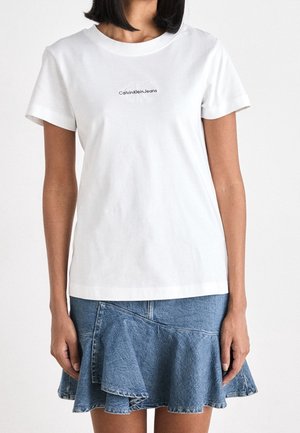 Nainen pukeutuneena valkoiseen Calvin Klein Jeans -T-paitaan ja siniseen farkkuhelmaiseen hameeseen seisoo vaaleaa yksiväristä taustaa vasten.