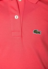 Pikepaita kirkkaan vaaleanpunaisena, jossa on teksturoidusta kankaasta valmistettu pinta. Ominaisuuksiin kuuluu klassinen kaulus, kolme valkoista nappia ja vihreä brodattu krokotiililogo.