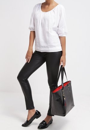 Una modella indossa una camicia bianca e pantaloni neri, tenendo una borsa tote nera con un interno rosso. Porta dei mocassini neri lucidi.