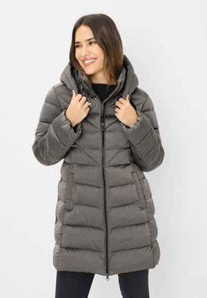Frau lächelnd, trägt langen grauen gepolsterten Wintermantel mit Kapuze, hält den Kragen mit den Händen, schwarze Hose, einfarbiger weißer Hintergrund.