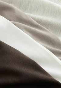 Lähikuva pehmeästä kankaasta, jossa on kerrokselliset vaakaraitoja tummanruskeassa, valkoisessa, vaaleanruskeassa ja kerma sävyissä.