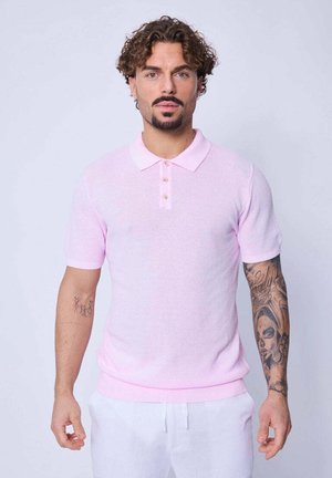 Homme aux cheveux bouclés et à la barbiche, portant un polo à manches courtes rose clair et un pantalon blanc, montrant des tatouages détaillés sur son bras droit.