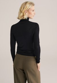 Juodas ilgarankovis golfo megztinis iš minkštos medžiagos, su prisitaikančia forma ir raukinėtomis rankovėmis, derinamas su alyvuogių spalvos pritaikytomis kelnėmis.