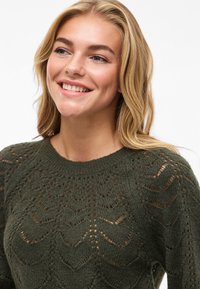 Χαμογελαστή γυναίκα με ξανθά μαλλιά, ντυμένη με σκούρο πράσινο πλεκτό πουλόβερ με σχέδια όπως δαντέλα, φόντο σε λευκό χρώμα.