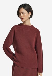 Burgundy stickad tröja med hög hals, ribbad textur och långa ärmar. Har en avslappnad passform och subtila sömdetaljer.