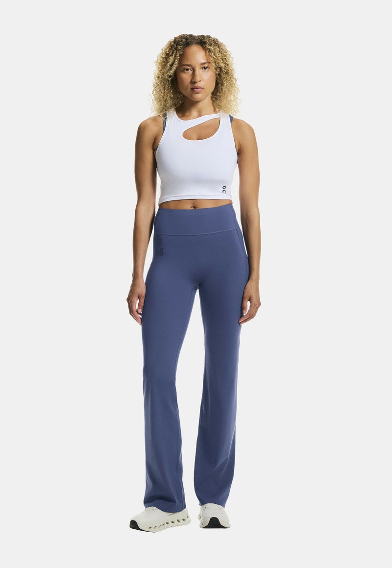 Biały sportowy crop top z wycięciem, zestawiony z niebieskimi spodniami o wysokim stanie, noszony w białych sneakersach. Dopasowany krój, gładka tekstura.