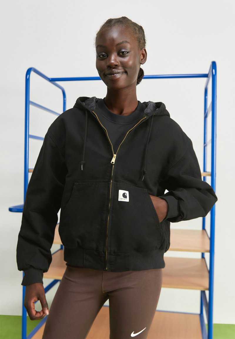 Black zip-up hoodie with a textured finish, gold zipper, and front pocket; worn over a black shirt, paired with brown leggings.