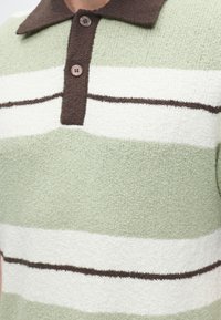Gros plan d'un polo en maille avec des rayures horizontales vert clair, blanc et marron et deux boutons marron sur la patte de col.