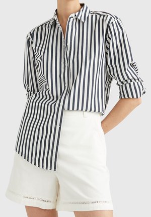 Gestreiftes Hemd in Marineblau und Weiß, mit Kragen und aufgerollten Ärmeln; kombiniert mit weißen Shorts mit dekorativem Saum.