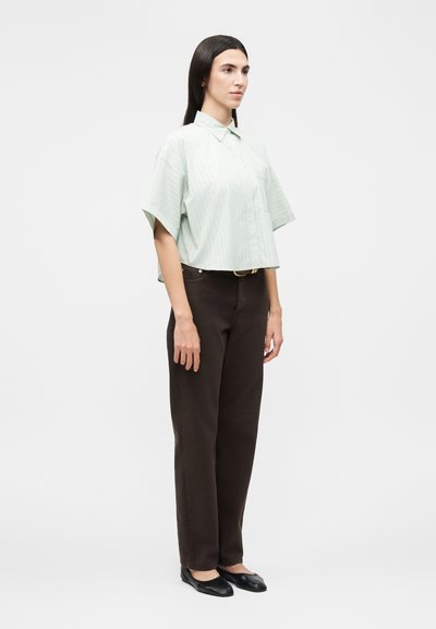 Νέος άνθρωπος στέκεται μπροστά φορώντας ένα φαρδύ ριγέ πουκάμισο με κοντά μανίκια, σκούρο ίσιο παντελόνι και μαύρα επίπεδα παπούτσια σε απλό φόντο.