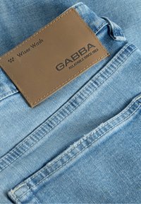 Tela de mezclilla azul claro con una textura suave, que presenta un parche de cuero marrón etiquetado "GABBA" y "Wiser Wash". Detalles de costura visibles.