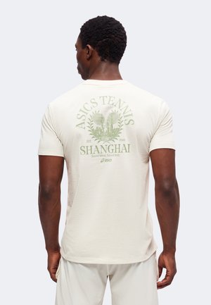 Άνδρας που φοράει φωτεινό T-shirt με το "ASICS TENNIS SHANGHAI" και γραφικό θέμα τένις στην πλάτη, σε συνδυασμό με ανοιχτόχρωμο σορτς.