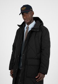 Černá prošívaná parka s kapucí, včetně vertikálního a horizontálního prošití, přední kapsy, nošená přes světle modrou košili a vzorovanou kravatou.