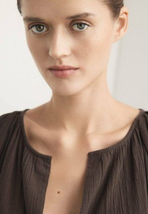 Giovane donna con occhi verdi e pelle chiara che indossa una camicetta marrone scuro con trama e scollatura a barca, espressione neutra, sfondo chiaro.