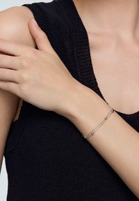 Srebrna bransoletka łańcuszkowa z dwoma sferycznymi akcentami, noszona na nadgarstku. W tle widać teksturowany czarny top dzianinowy.