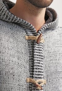 Primo piano di un uomo che indossa un maglione a collo alto grigio in maglia con due chiusure a toggle in legno fissate con anelli di corda beige.