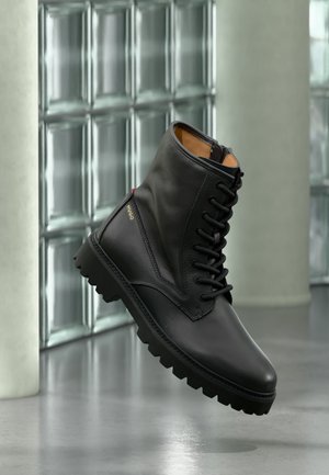 Black leather lace-up combat boot with thick sole, gold "HUGO" logo on side, displayed in modern indoor setting with glass block wall background.