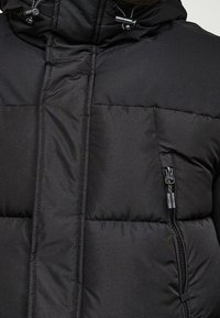 Černá prošívaná bunda s kapsou na zip. Disponuje hladkou, texturovanou látkou a vysokým límcem s nastavitelnými stahovacími šňůrkami na kapuci.