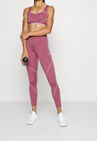 Completo di reggiseno sportivo e leggings color marsala con cuciture riflettenti bianche, abbinato a sneakers bianche. La modella tiene in mano una bottiglia d'acqua nera.
