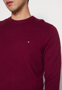 Burgunderfarbener Strickpullover mit rundem Halsausschnitt, gerippten Bündchen und Saum. Verfügt über ein kleines Logo mit blauen, weißen und roten Akzenten auf der Brust.