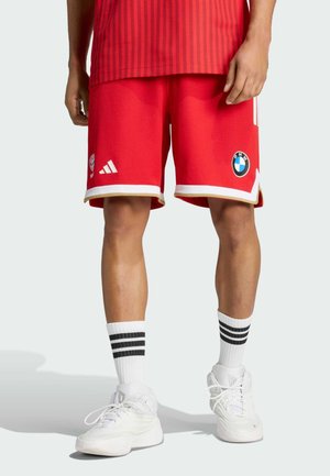 Osoba ubrana w czerwone sportowe spodenki Adidas z logo BMW i drużyny, białe skarpetki z czarnymi paskami oraz białe buty sportowe, stojąca na jasnej podłodze.