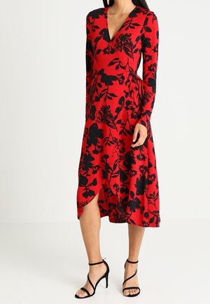Femme portant une robe portefeuille rouge avec un imprimé floral noir et des sandales à talons noirs, debout sur un fond blanc uni.