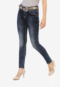 Blaue Skinny-Jeans mit einem verwaschenen Design, ausgestattet mit Seitentaschen und goldfarbenen Beschlägen, kombiniert mit beigen High-Heels.
