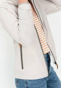 Personne portant une veste beige sur une chemise à rayures orange et un jean bleu, ajustant le col de la veste avec la main gauche.