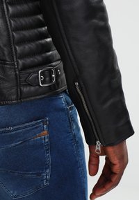 Svart läderjacka med quiltade axeldetaljer, silverfärgad dragkedja på ärmen och ett spänne vid fållen, tillsammans med blå jeans.