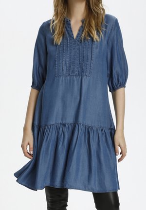 Kobieta ubrana w luźną niebieską sukienkę z denimu z detalami plisowania, rękawami do łokcia i marszczoną spódnicą, w połączeniu z czarnymi spodniami.