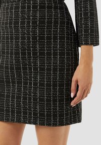 Schwarzes Kleidungsmaterial mit strukturierter Oberfläche, das ein weißes Rautenmuster aufweist. Kurzarmdesign, das knapp über dem Knie endet, mit einer figurschmeichelnden Silhouette.