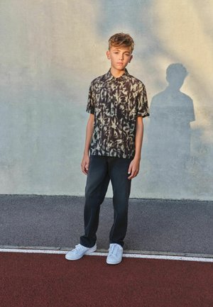 Teismeline poiss lühikeste lokkis juustega seisab tänavalävel seina kõrval, kandes mustriga lühikese varrukaga särki, musti pükse ja valgeid vabaajajalatsid.